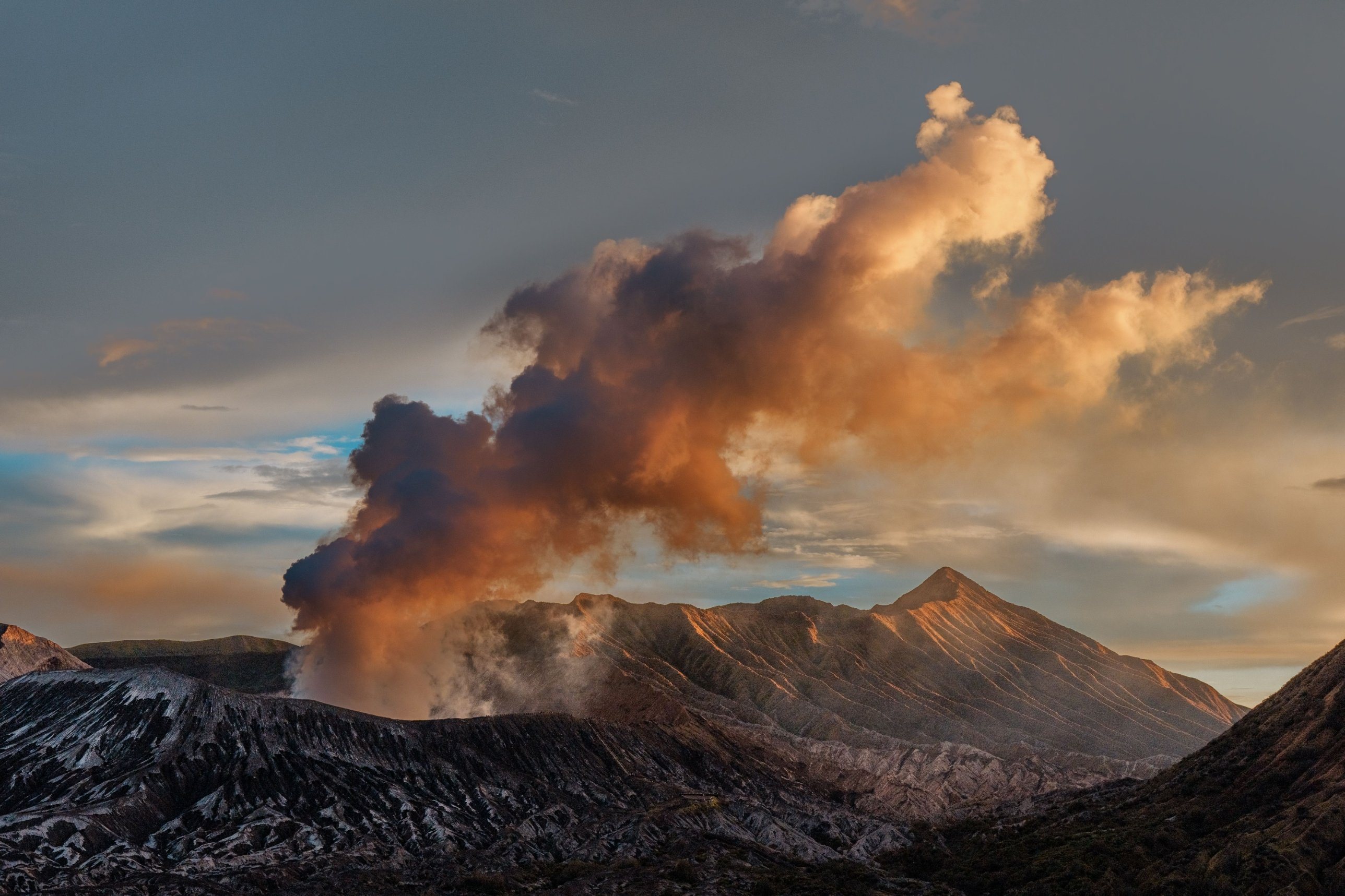 Mount Bromo sending a plume of ash into the evening sky, with Mount Batok beside it